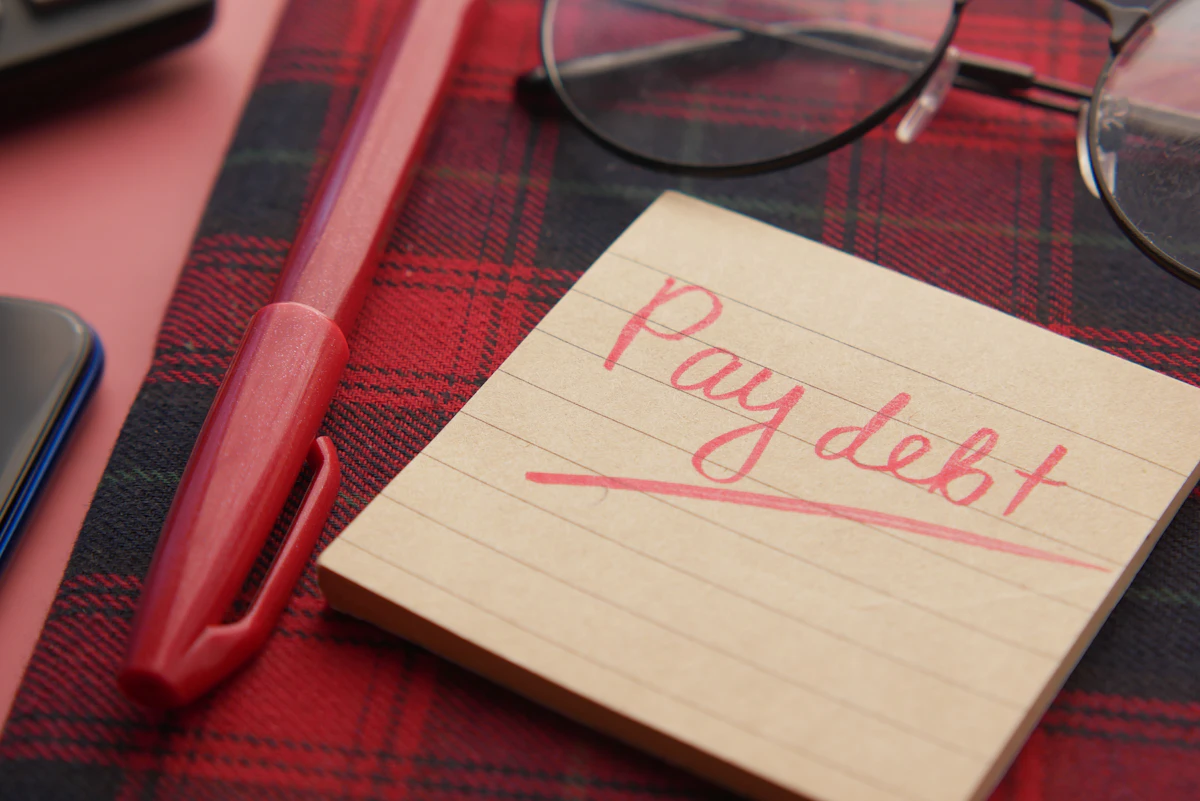 Note reading pay debt next to a pen and glasses
