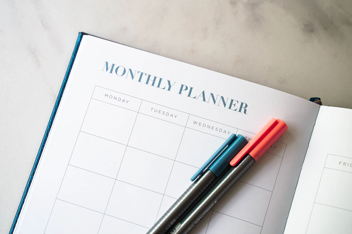 Open monthly planner with teal and coral pens on marble desk